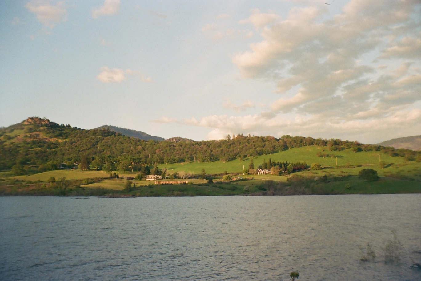 A photo of a green hill and trees and houses on the other shore of a lake