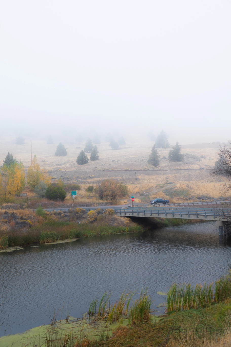 A photo of a truck driving down a road by a river on a foggy morning