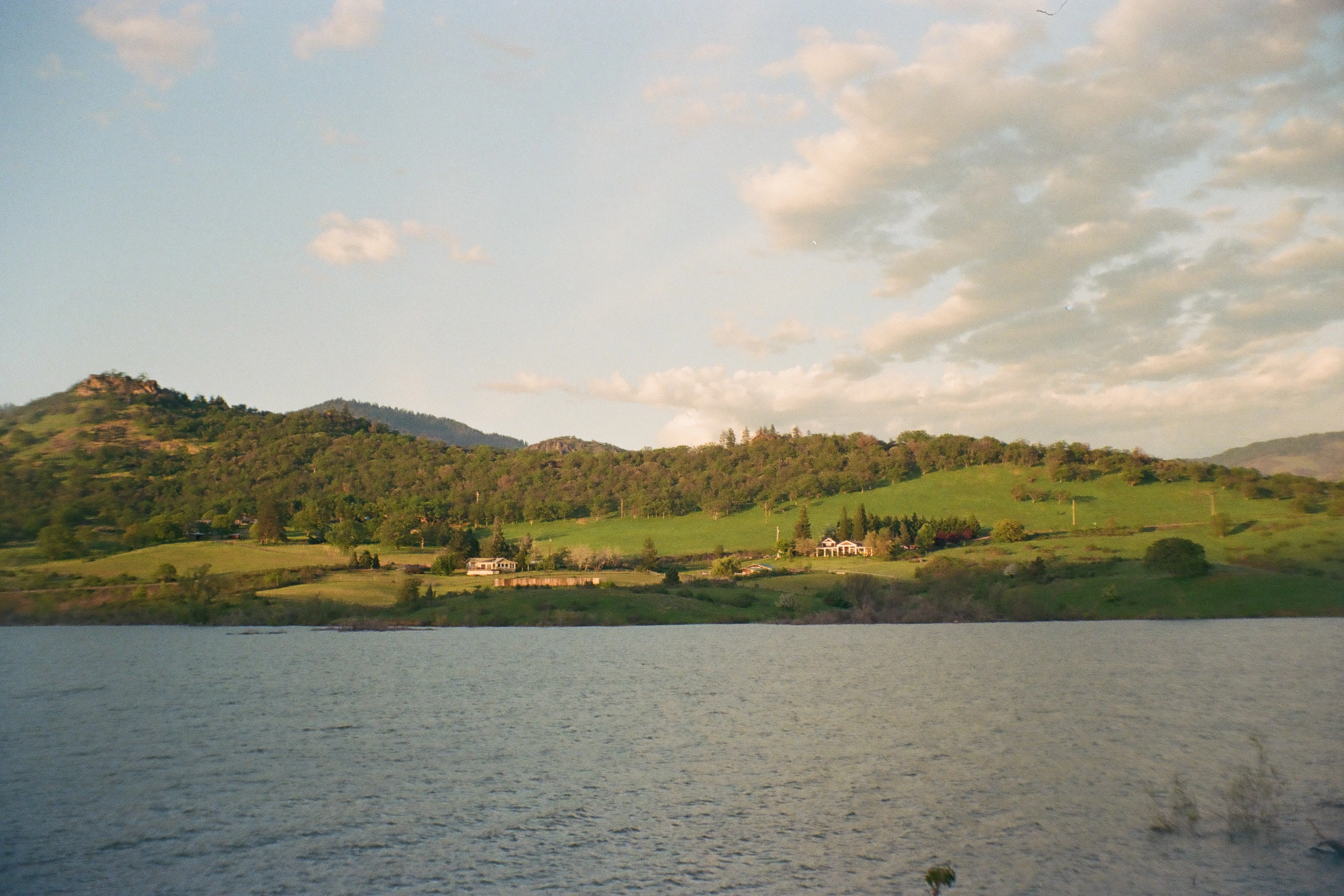 A photo of a green hill and trees and houses on the other shore of a lake